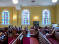 A view of the back right and middle section of the sanctuary, looking from the left side of the sanctuary across to the right side of the sanctuary. Three of the four domed stained-glass windows on the right wall of the sanctuary are visible. The square stained-glass window on the right side of the back wall is partially visible on the right side of the photograph. The covered table in the back, right corner of the sanctuary is partially visible between the first domed stained-glass window and the square stained-glass window. The glass-front bookcase is partially visible between the first and second domed stained-glass windows.