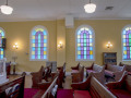 A view of the front right and middle sections of the sanctuary. Three of the domed stained-glass windows are visible on the right wall of the sanctuary. The fourth domed stained-glass window on the right wall of the sanctuary is partially visible on the left side of the photograph. The glass-front bookcase on the right wall of the sanctuary is partially visible between the first and second domed stained-glass windows. The red carpeted area separates the middle section of the sanctuary from the right side of the sanctuary. The white rail enclosing the front of the bema is partially visible on the left side of the photograph. A small table and menorah are visible between the third and fourth domed stained=glass windows. The pews on the right side of the bema are partially visible to the left of the small table and menorah.