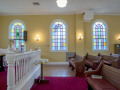 A view of the front and middle pews, looking from the left side of the sanctuary across to the right side of the sanctuary. The glass-front bookcase is partially visible on the right side of the photograph. Three of the four domed stained-glass windows on the right wall of the sanctuary are visible or partially visible. The white rail across the front of the bema and a small table outside the bema are visible. The large pulpit is partially visible on the left side of the photograph.
