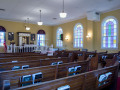 A view of the sanctuary, looking from the back, left corner of the room toward the front, right corner of the room. The open ark, and the framed artwork and domed windows on either side of it are visible. The front and left side of the white rail enclosing the bema is partially visible. The two pews to the right of the bema, facing the bema, and the standing menorah are partially visible. Three of the four domed, stained-glass windows are visible on the right side of the sanctuary. The glass-front bookcase on the right wall of the sanctuary is partially visible on the right side of the photograph.