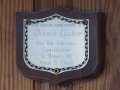 A close-up view of the three plaques hanging on the wall of the long side of the L-shaped area, under the ceiling overhang, on the left, front side of the social hall. The top plaque (Dinnie Lusher)