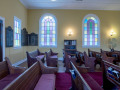 A view of the back section of the sanctuary, looking from the right side of the room across to the left side of the room. The left side of the front doors is partially visible on the left side of the photograph. To the left of the front door is a large yahrzeit plaque. To the left of that yahrzeit plaque is the square, stained-glass window on the front, left side of the building. To the left of the square, stained-glass window is another large yahrzeit plaque. Two domed, stained-glass windows and part of a third domed, stained-glass window are visible on the left wall of the sanctuary. A small yahrzeit plaque is to the left of the first domed, stained-glass window. A glass-front bookcase is partially visible between two of the domed, stained-glass windows on the left wall of the sanctuary. The Tree of Life is partially visible to the left of the second domed, stained-glass window.
