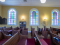 A view of the back and middle sections of the sanctuary, looking from the right side of the room across to the left side of the room. Three of the four domed, stained-glass windows on the left wall of the building are visible. The large yahrzeit plaque on the back, left wall and the smaller yahrzeit plaque to the left of it are visible. The glass-front bookcase on the left wall is partially visible between the first and second domed, stained-glass windows. The Tree of Life, on the left wall, is partially visible between the second and third domed, stained-glass windows. The red carpeted area separates the wooden floored are on the left side of the sanctuary from the middle section of the sanctuary.