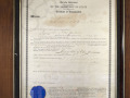 A close-up view of the framed document (also a Certificate of Incorporation) hanging on the back wall, above and to the right of the glass-front cabinet.