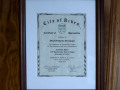 A close-up view of the framed certificate hanging to the left of the grey door on the far, left end of the left wall of the social hall.