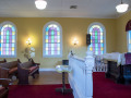 A view of the front area of the sanctuary, looking from the right side of the room across to the left side of the room. Three of the four domed, stained-glass windows on the left side of the sanctuary are visible. The Tree of Life is partially visible between the second and third domed, stained-glass windows. The white rail across the front of the bema and the small table in front of it are visible. The white rail on the left side of the bema is partially visible below the third, domed stained-glass window. The front and right side of the pulpit are visible on the bema. The domed, stained-glass window to he left of the ark is partially visible behind the American flag.
