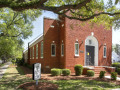 A view of the front and left side of the synagogue, looking from the corner of Screven and Highmarket Streets toward the synagogue. The sign with the synagogue name and information is visible on the lawn, on the front, left side of the building. The cornerstone of the building is visible between the bushes on the left side of the building and the sign on the front lawn. A metal plaque commemorating the founding and dedication of the synagogue is visible on the front of the building, to the left of the front doors. A square, stained glass window and commemorative plaque are inset in the white arch above the front doors. The stained glass windows on each side of the front doors and two stained glass windows on the front, left side of the sanctuary are visible. The partially visible, large windows on the left side of the building are stained glass windows in the sanctuary. The round, stained glass Star of David window toward the top of the front of the building is not visible in this photograph.