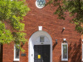 A close-up view of the front of the synagogue. A metal plaque commemorating the founding and dedication of the synagogue is attached to the bricks to the left of the front doors. A square, stained glass window and commemoration plaque are inset in the white arch above the front doors. A round, stained glass window with the Star of David on it is visible toward the top of the building. A stained glass window is on each side of the front doors.