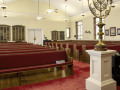 A view of the sanctuary, looking from the front, left corner of the sanctuary toward the back, right corner of the sanctuary. The bema is partially visible on the far, right side of the photograph. The double doors on the back wall of the sanctuary divide the front entry area to the synagogue from the sanctuary.