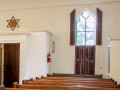 A close-up view of the back wall on the right side of the sanctuary. The open door to the entry way is visible on the left side of the photograph. A glass-front cabinet is partially visible on the right side of the photograph. The window is the left, front window of the synagogue.
