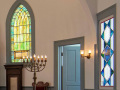 A view of the left, front corner of the sanctuary, showing one of the green stained-glass windows, one of the standing menorahs, and the first stained-glass window on the left side of the sanctuary. The blue door between the stained-glass windows leads to a small room.