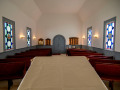 A view of the sanctuary, looking from the bema toward the front doors of the building. Two of the three stained-glass windows on both the left and right sides of the sanctuary are partially visible. Three yahrzeit plaques are visible on the wall by the front doors. A small table with literature about the synagogue is partially visible in the back, right corner of the sanctuary.