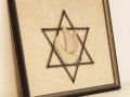 A close-up view of the framed, artwork on the empty bookcase at the entrance to the Social Hall, showing an embroidered dove within a Star of David. The Star of David is made of metal tacks.